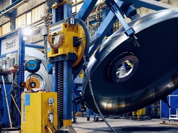 A worker wearing protective clothing and a blue helmet operates machinery on a large metal component in an industrial workshop. The background shows equipment and tools.