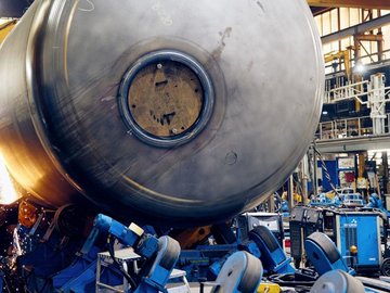 Large cylindrical metal tank being processed in a manufacturing hall, with sparks flying during welding